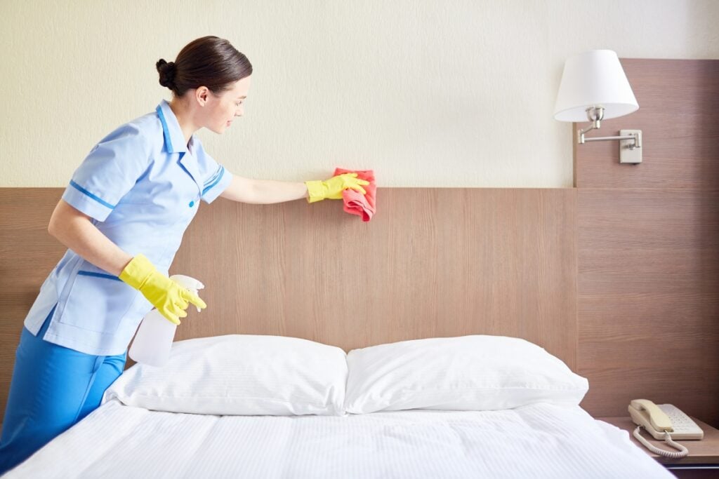 Staff dusting bed with sponge in hotel-room.