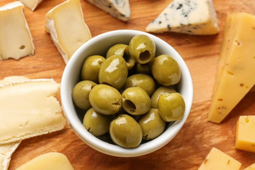 Olives in a bowl with assorted cheeses on a wooden board.