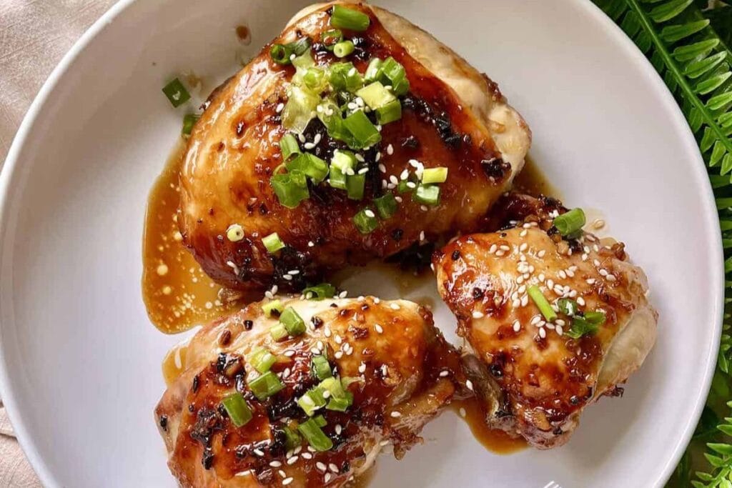 Three glazed chicken thighs garnished with sesame seeds and green onions on a white plate.