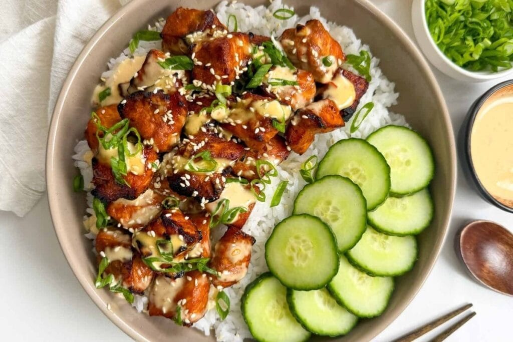 Salmon bites over white rice with sliced cucumbers, drizzled with sauce and scallions.