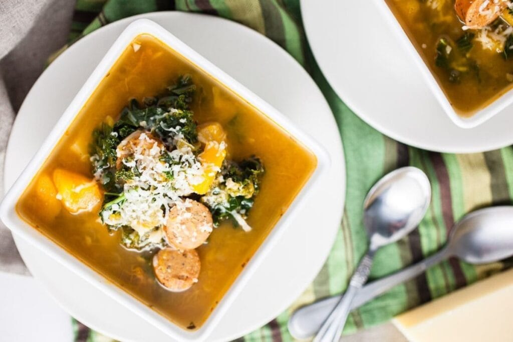 Butternut squash and sausage soup in a white bowl, with spoons and cheese.