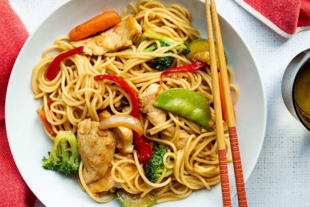 A white bowl of chicken and vegetable lo mein with chopsticks resting on the side.