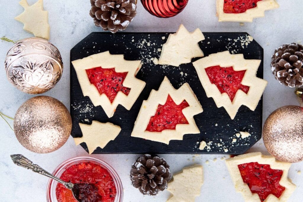 Christmas tree linzer cookies with red filling on a black board, surrounded by ornaments.