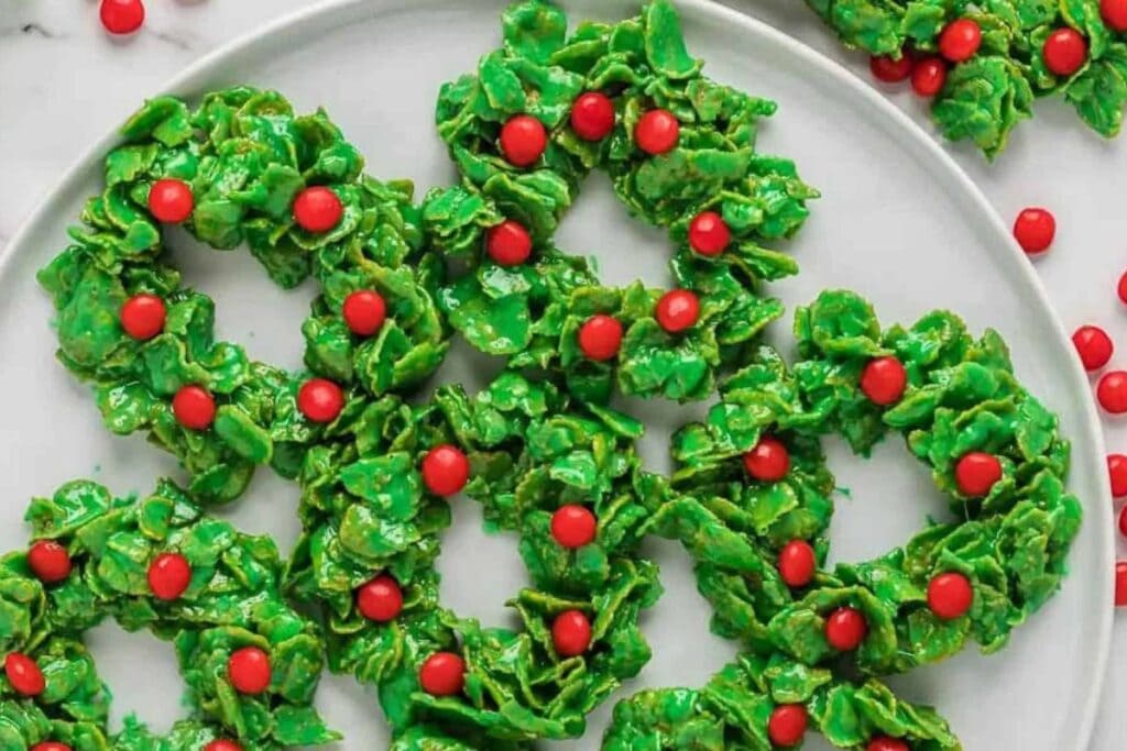 Green cornflake wreath cookies with red candy "berries" on a white plate.
