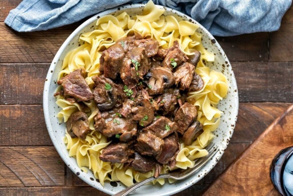 Beef tips and mushrooms over egg noodles in a bowl, garnished with parsley.