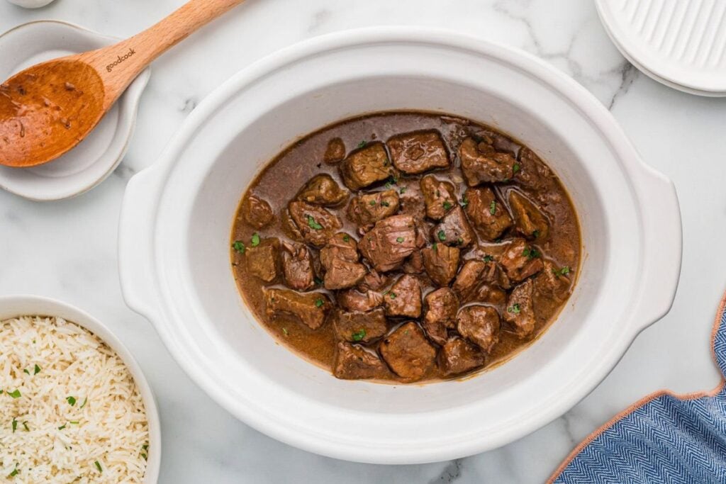 Honey bourbon steak in a white slow cooker with rice and a wooden spoon.