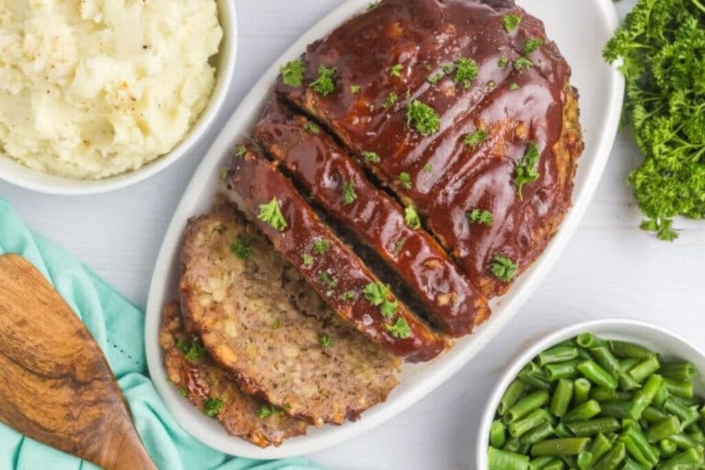 Meatloaf with BBQ sauce, mashed potatoes, and green beans on a white platter.