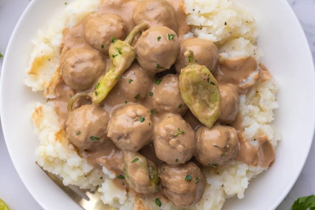Meatballs with gravy and pepperoncini peppers over mashed potatoes on a plate.