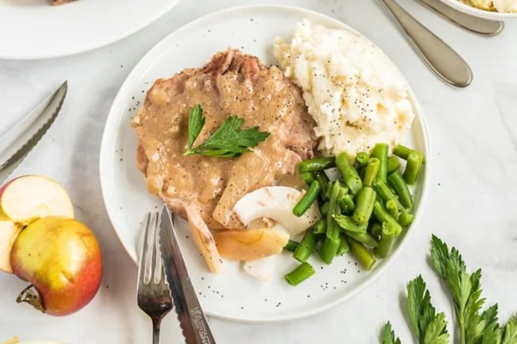 Pork chop with gravy, mashed potatoes, green beans, and cooked apples on a plate.