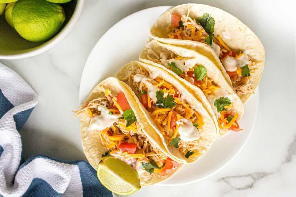 Four chicken tacos on a white plate, topped with cheese, tomatoes, and cilantro.