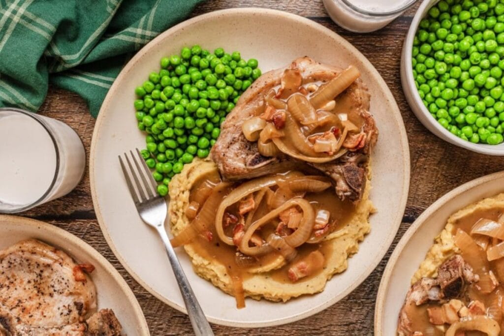 Pork chop with gravy and onions, mashed potatoes, and peas on a plate.