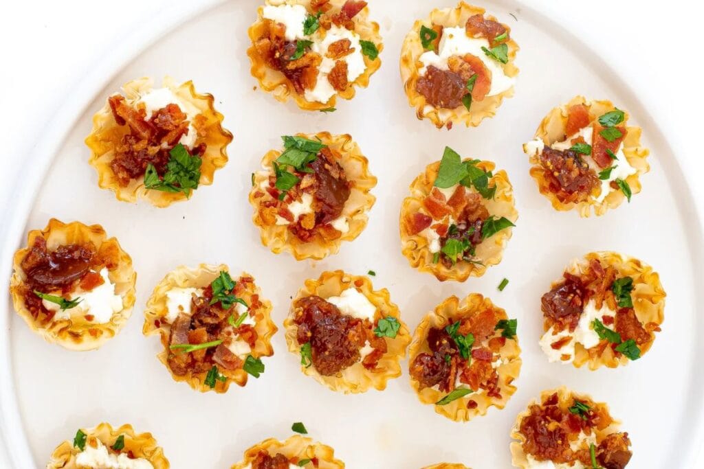 Phyllo cups filled with goat cheese and fig jam topped with bacon and parsley on a white plate.