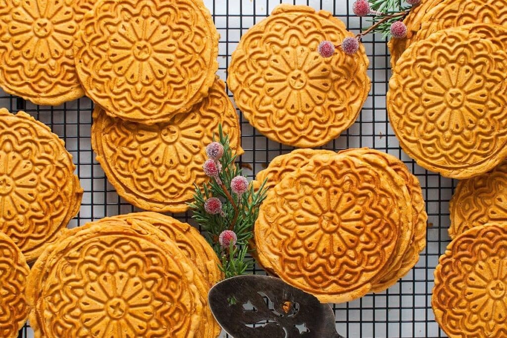 Overhead shot of golden brown pizzelle cookies on a cooling rack with holiday greenery.