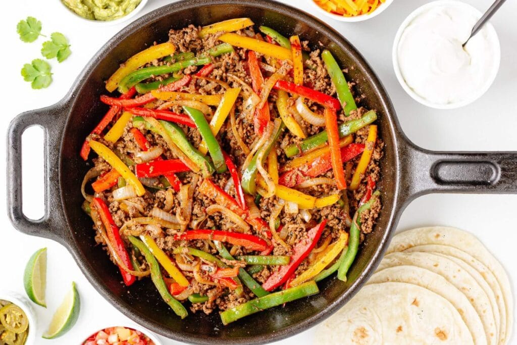 Ground beef fajitas with peppers and onions in a skillet, with tortillas and toppings.