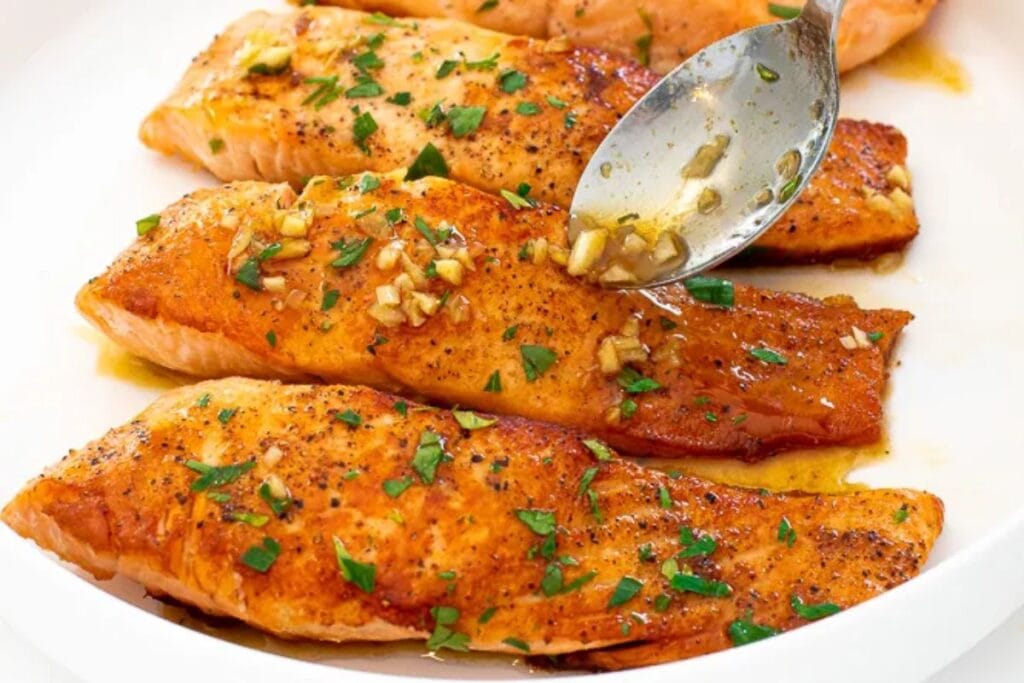 Baked salmon fillets being drizzled with a garlic butter sauce.
