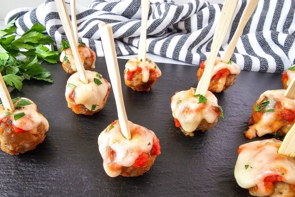 Meatballs with marinara sauce and melted cheese on skewers, served on a black slate.