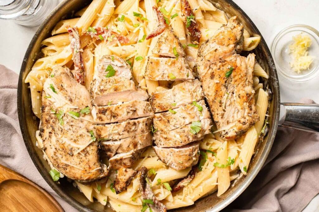 Penne pasta with sliced chicken breast, parmesan cheese, and herbs in a skillet.