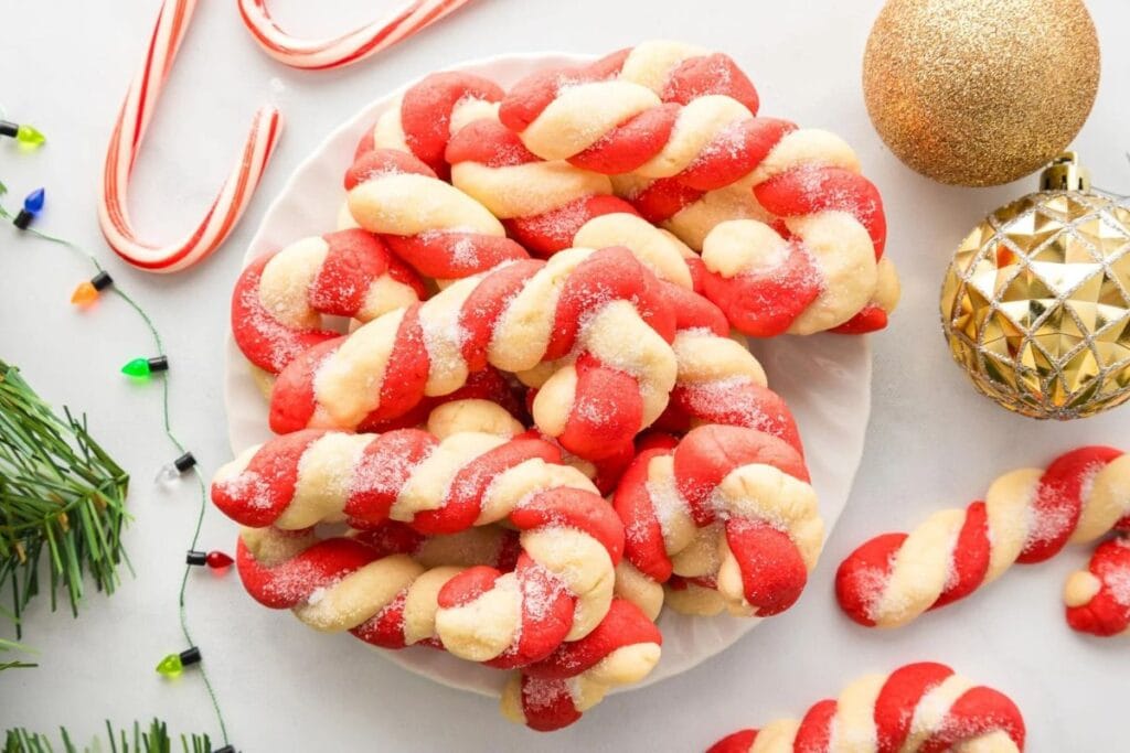 Candy cane-shaped cookies, red and white twisted together, sprinkled with sugar on a white plate.