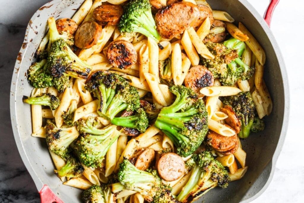 Penne pasta, sliced sausage, and charred broccoli florets in a gray skillet.