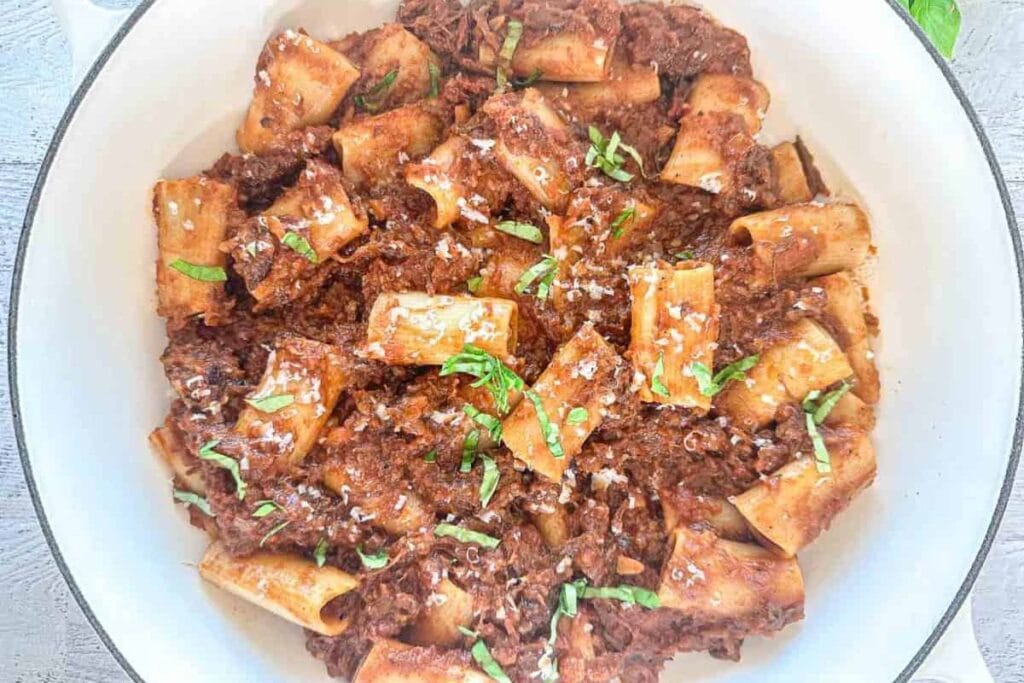 Rigatoni pasta with short rib ragu, parmesan cheese, and basil in a white pot.