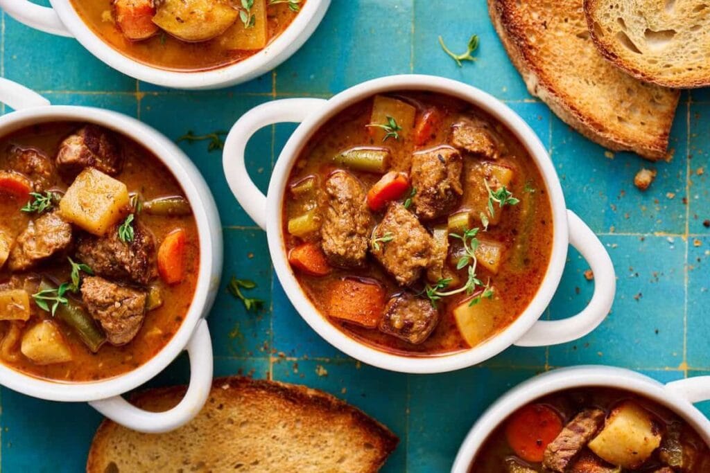 Overhead view of bowls of beef stew with toasted bread on blue tiles.