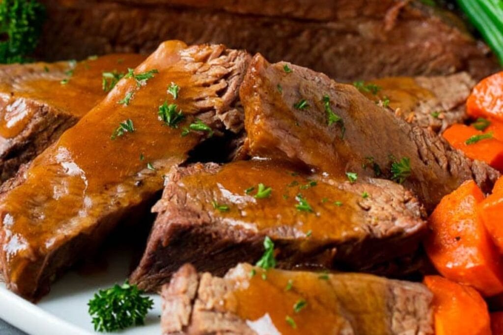 Sliced brisket with gravy, garnished with parsley, served with cooked carrots.