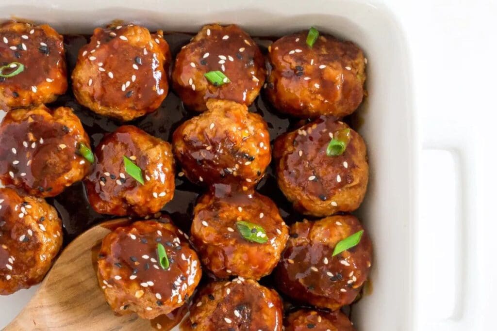 Meatballs with sriracha sauce, sesame seeds, and green onion garnish in a white dish.
