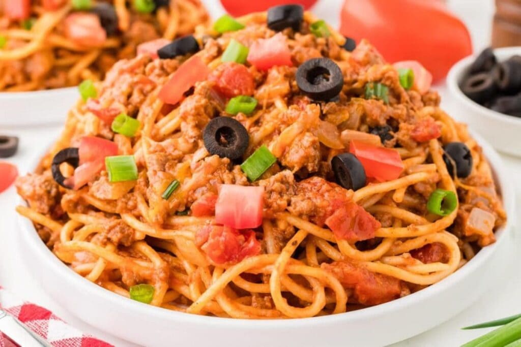 Close-up of taco spaghetti with tomatoes, olives, and green onions in a white bowl.