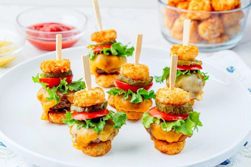 Mini cheeseburger bites with tater tot buns, lettuce, tomato, and pickle on a white plate.