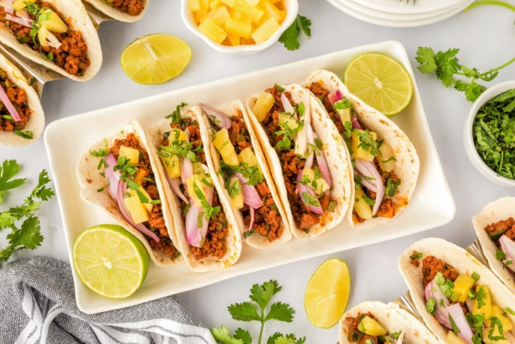 Al pastor tacos with pineapple, cilantro, and lime slices on a plate.