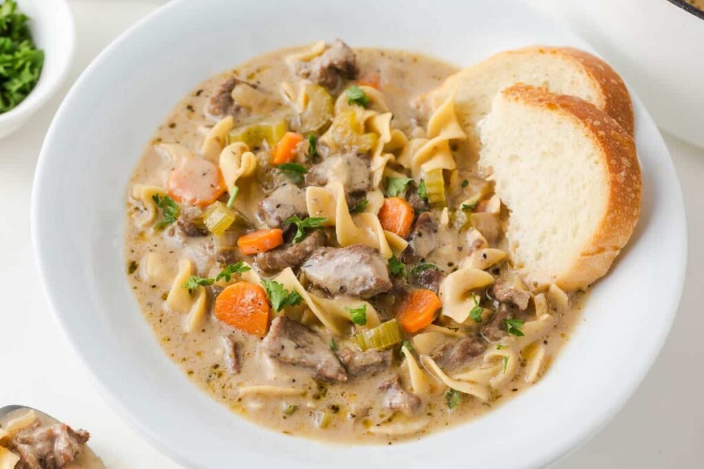 Beef noodle soup in a bowl with a slice of bread on the side.
