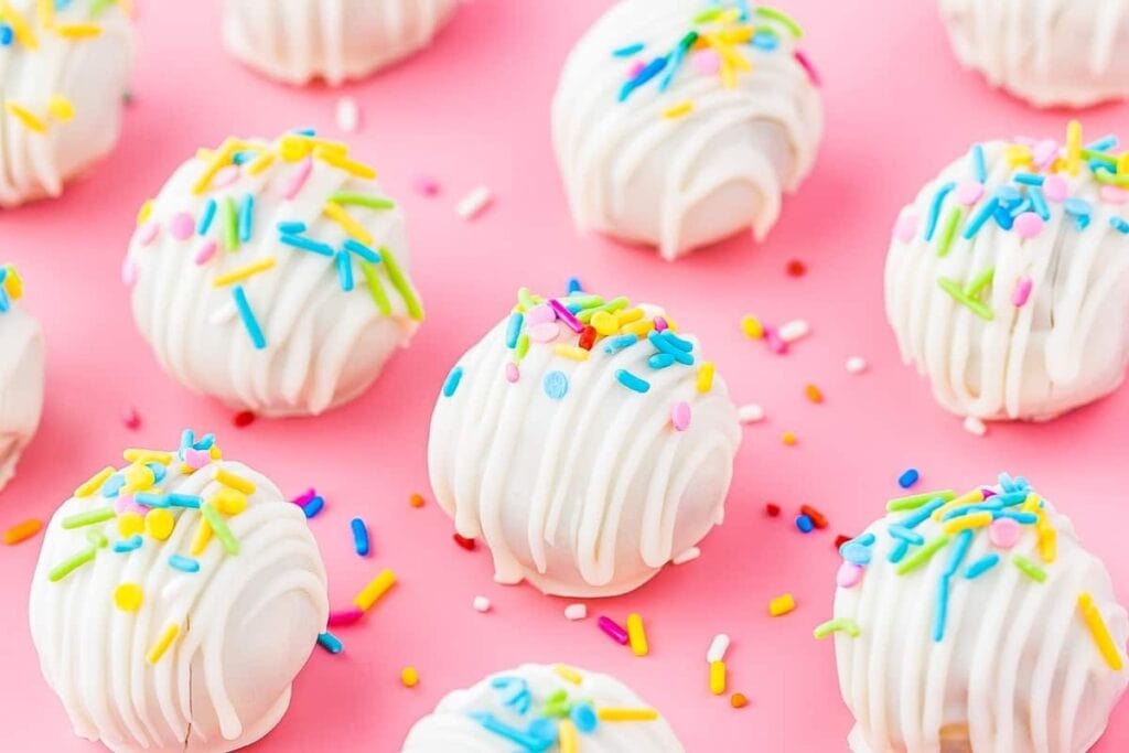 White cake pops with colorful sprinkles arranged on a pink surface.
