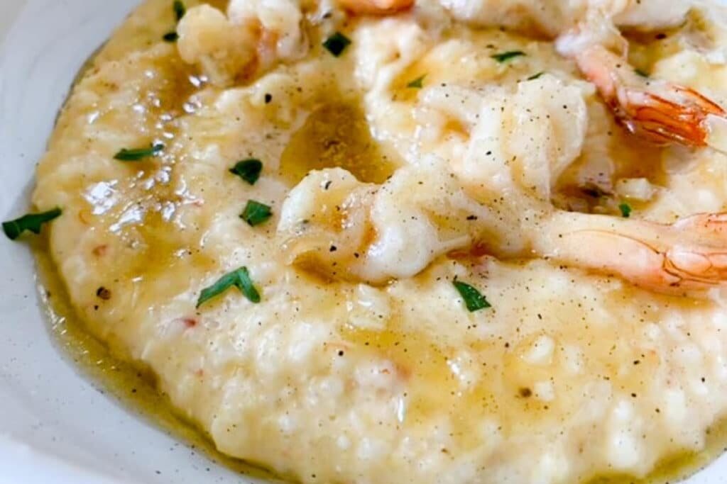  Close up of garlic butter shrimp served over creamy grits on a plate, garnished with herbs.