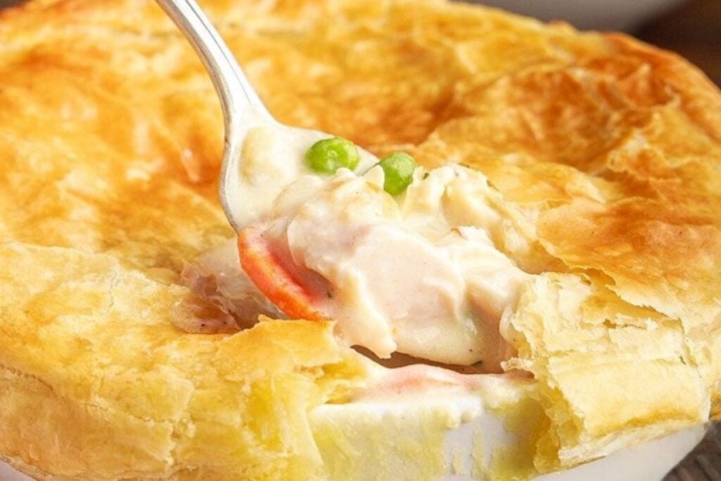 KFC chicken pot pie with a cracked golden crust in the center, showing creamy filling with vegetables on a spoon.