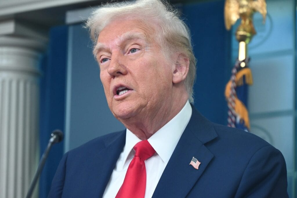 Trump in a suit and red tie speaking at a podium.