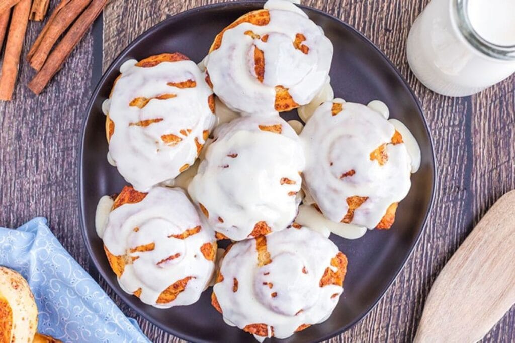 Cinnamon rolls with white icing on a black plate, with cinnamon sticks nearby.