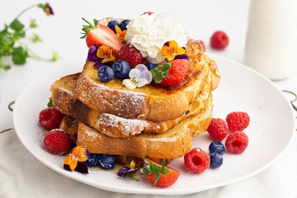 Stack of French toast topped with whipped cream, berries, and edible flowers.
