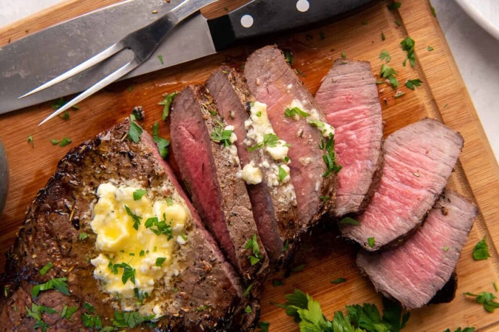 Sliced London broil steak with butter and parsley on a wooden cutting board.