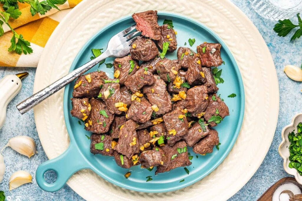 Overhead view of steak bites with garlic and parsley on a blue plate.