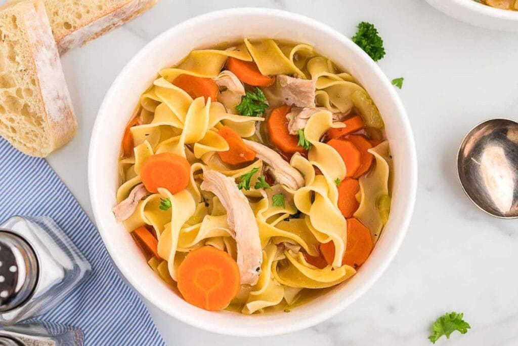 Overhead view of chicken noodle soup in a white bowl with bread and seasonings.