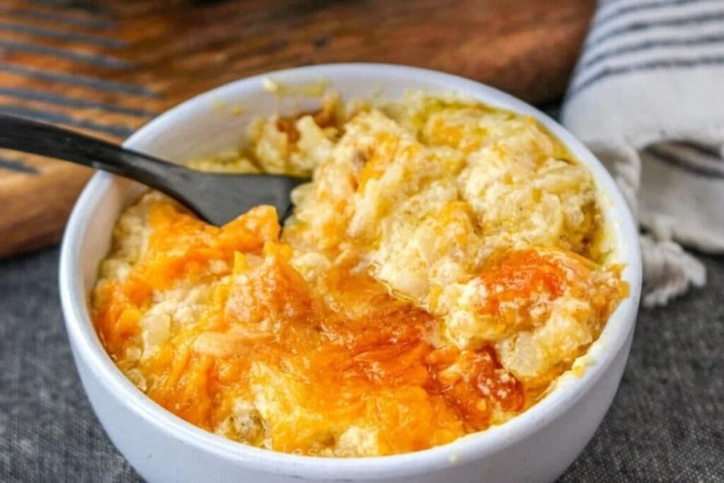 A bowl of cheesy hashbrown casserole with a spoon, on a gray surface.