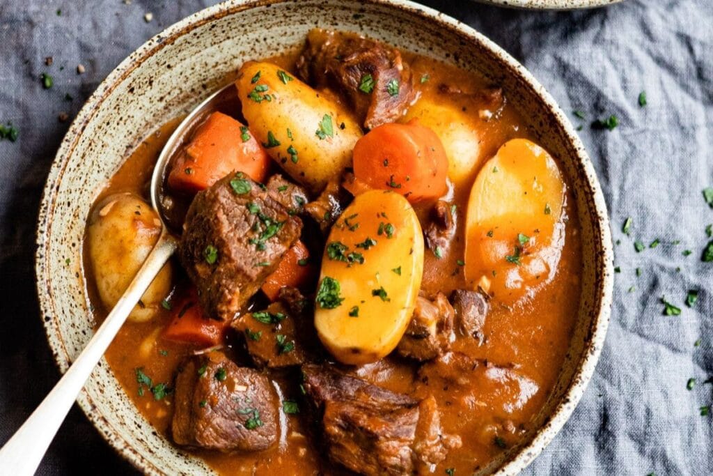 Close-up of beef stew with potatoes and carrots in a speckled bowl with a spoon.