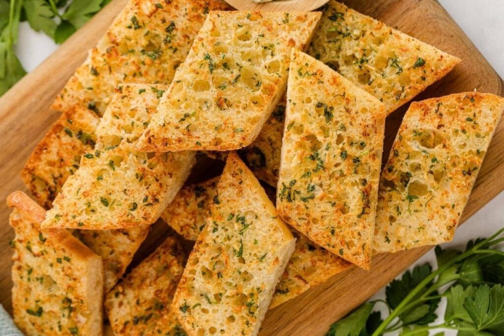 Garlic bread slices with herbs piled on a wooden cutting board.