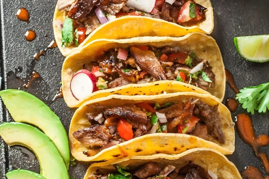 Overhead view of pork carnitas tacos with avocado and lime on a dark surface.