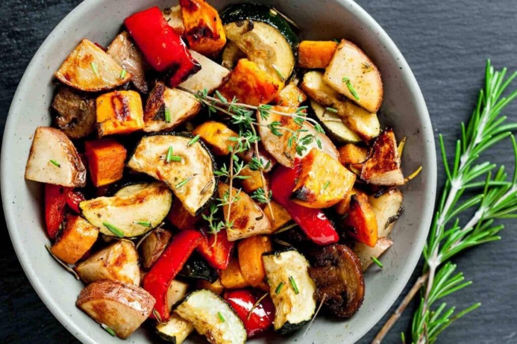 Roasted vegetables in a bowl with rosemary on a dark surface.