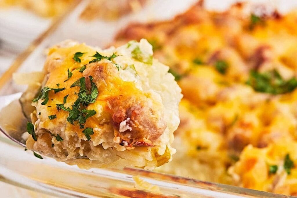 Spoon lifting a portion of sausage and hash brown casserole from a glass dish.
