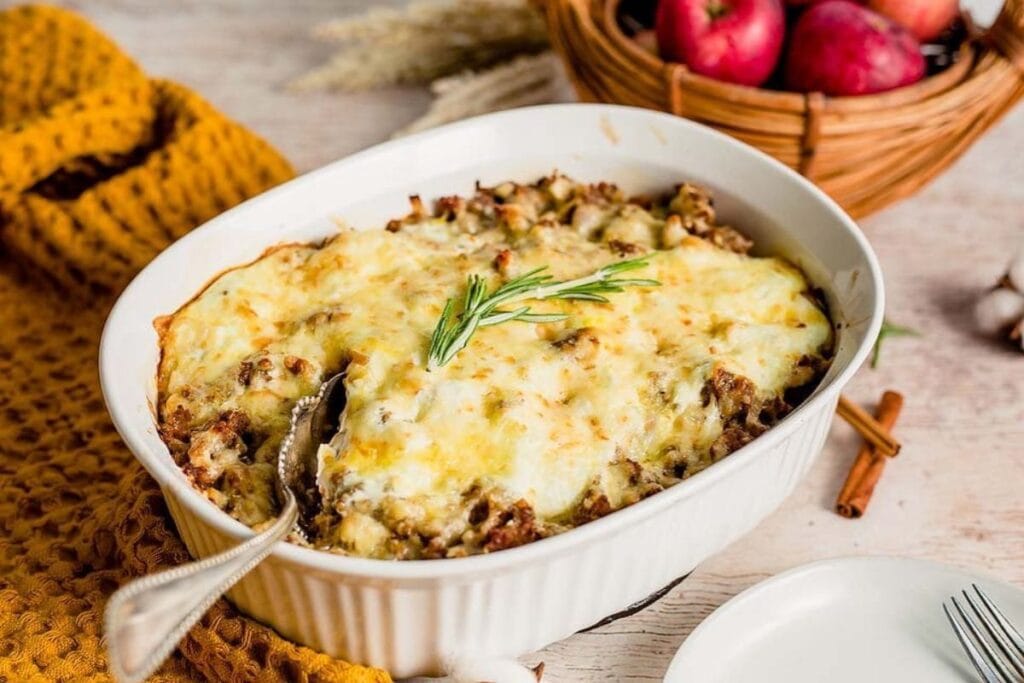 Sausage apple casserole with cheese in a white dish, garnished with rosemary.