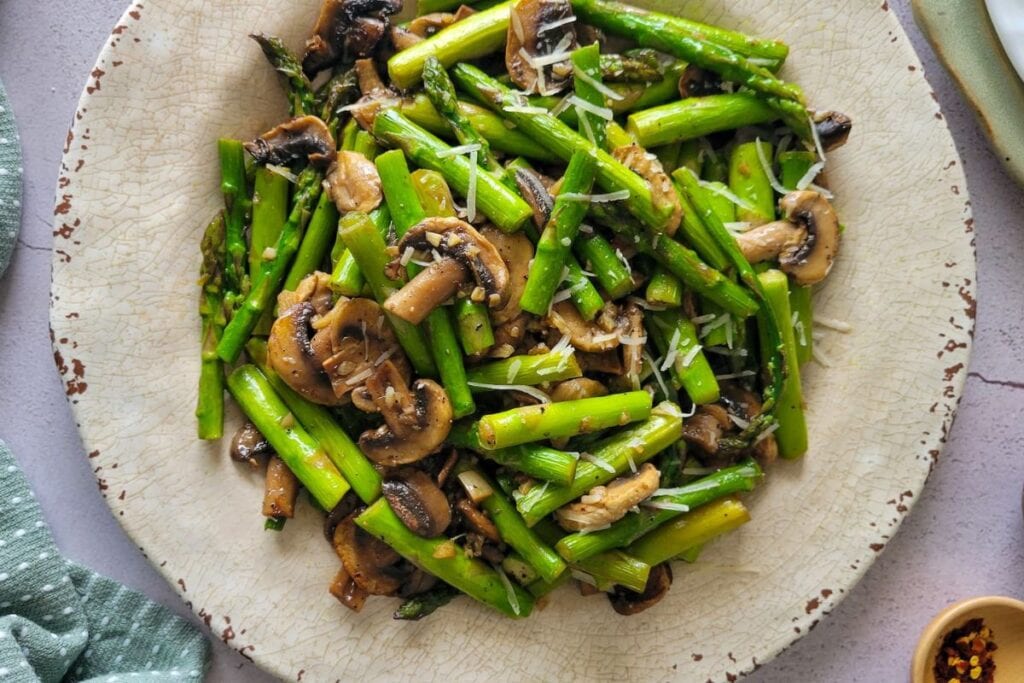Sautéed asparagus and mushrooms with parmesan on a speckled plate.