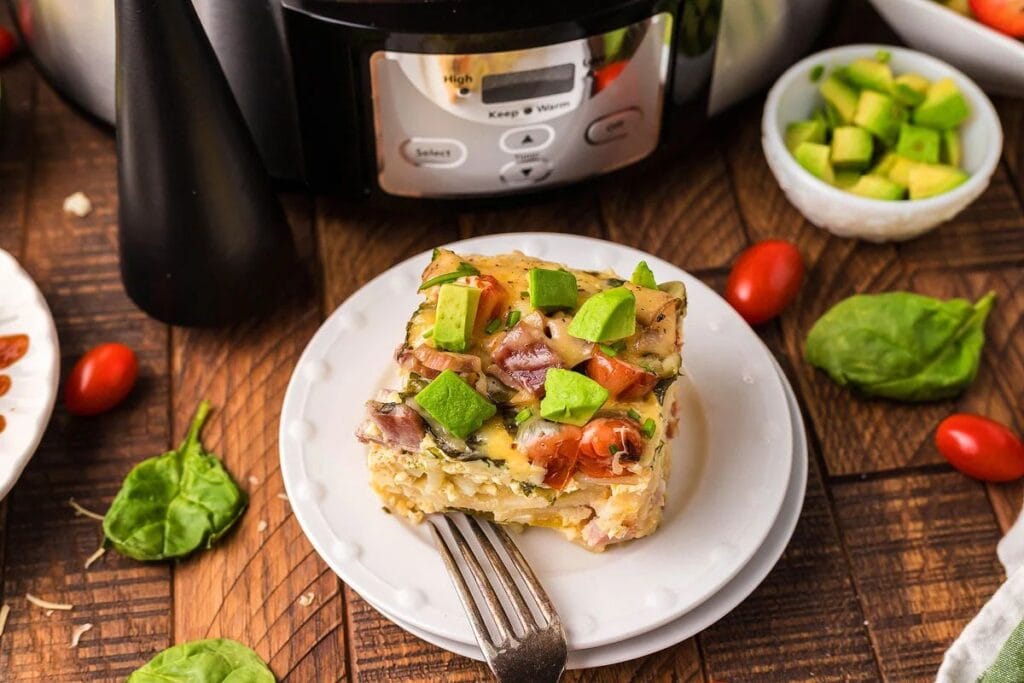A slice of breakfast casserole topped with avocado on a plate, with a slow cooker.