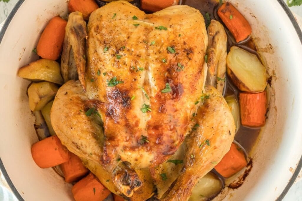 Whole roasted chicken in a Dutch oven surrounded by carrots and potatoes.